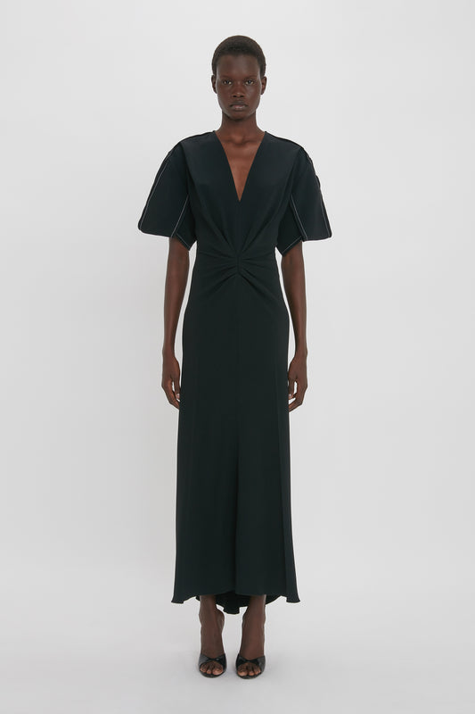 A person stands in a long, black Gathered V-Neck Midi Dress in Black by Victoria Beckham with short sleeves and a deep V-neck, made from figure-flattering stretch fabric, against a plain white background. They wear black high-heeled sandals, adding a contemporary edge to the look.
