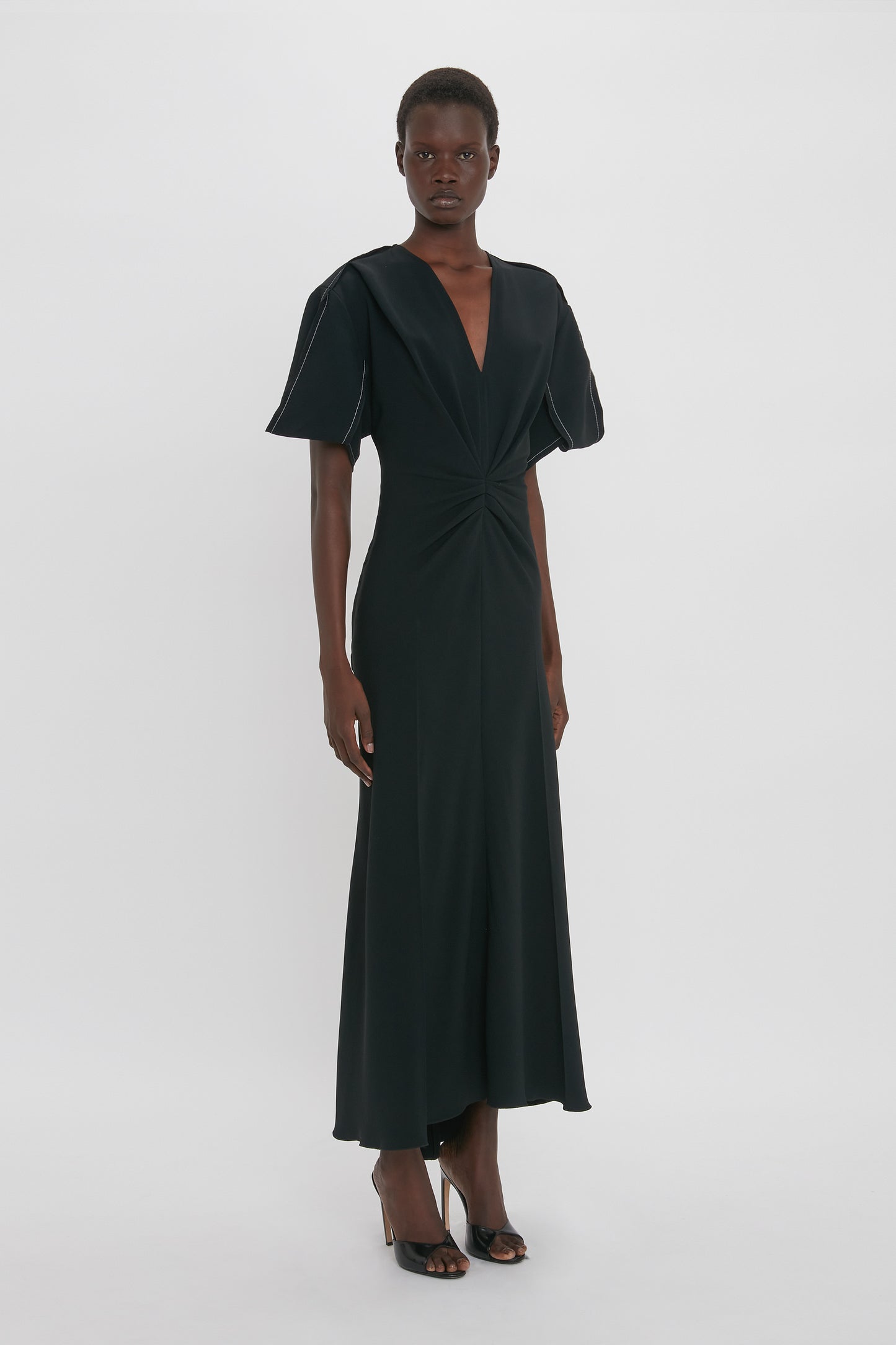 Person standing and modeling a Gathered V-Neck Midi Dress in Black by Victoria Beckham with short puffed sleeves and a gathered detail at the waist, crafted from figure-flattering stretch fabric for a contemporary edge. The dress is paired with black high-heeled shoes.