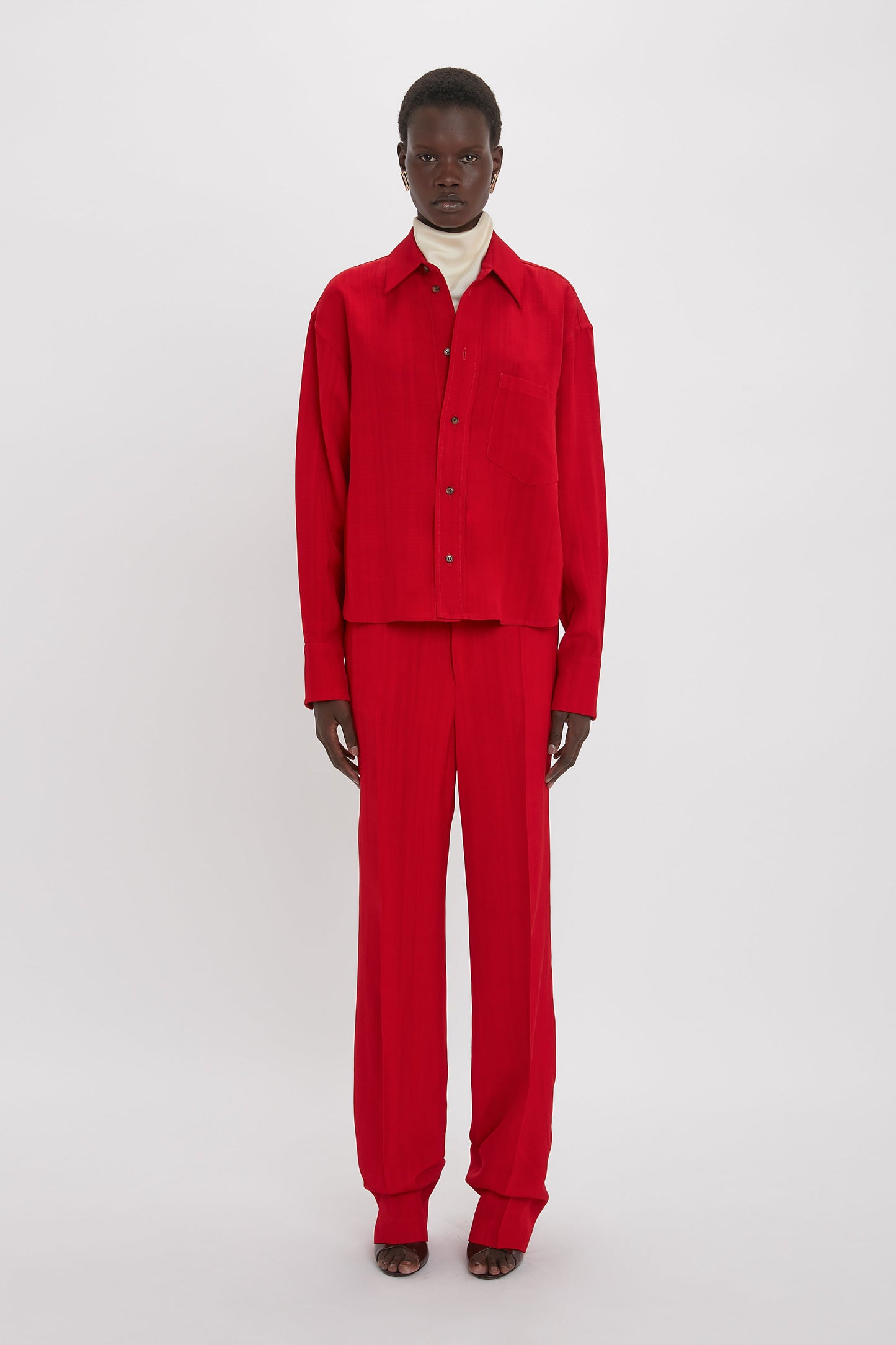 A person stands against a white background wearing a Victoria Beckham Cropped Long Sleeve Shirt In Carmine and matching red pants with a white turtleneck underneath, exuding a refined yet casual look.