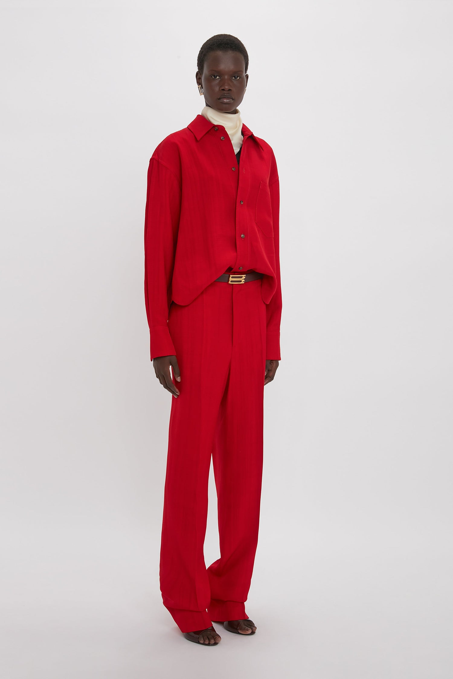 A person wearing a red shirt and matching Victoria Beckham Tapered Leg Trouser In Carmine stands against a plain white background, showcasing a lean silhouette that speaks to traditional tailoring.