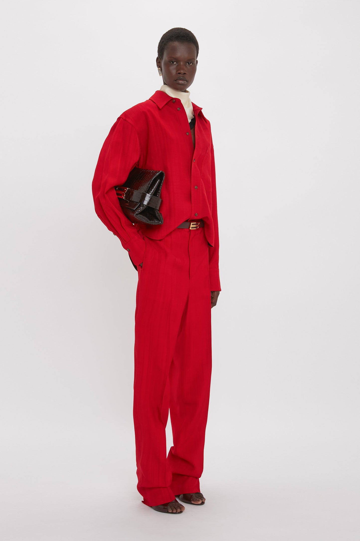 Person standing in a white space, wearing a red pantsuit with traditional tailoring and Tapered Leg Trouser In Carmine by Victoria Beckham, holding a black handbag.