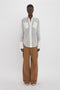 A person stands against a plain white background, exuding casual sophistication in a relaxed fit Pocket Detail Shirt In White by Victoria Beckham with large pockets and long sleeves, paired effortlessly with wide-leg brown pants and pointed white shoes.