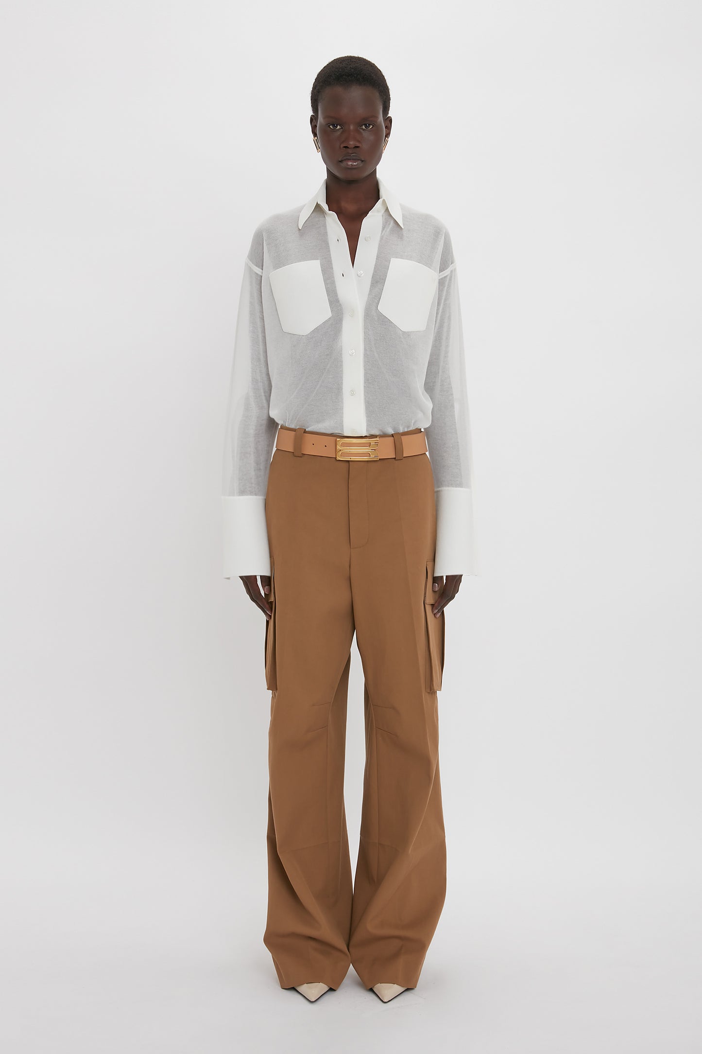 A person stands wearing a white Pocket Detail Shirt In White by Victoria Beckham with large pockets and wide brown pants, paired with a beige belt and pointed shoes, exuding casual sophistication against a plain white background.