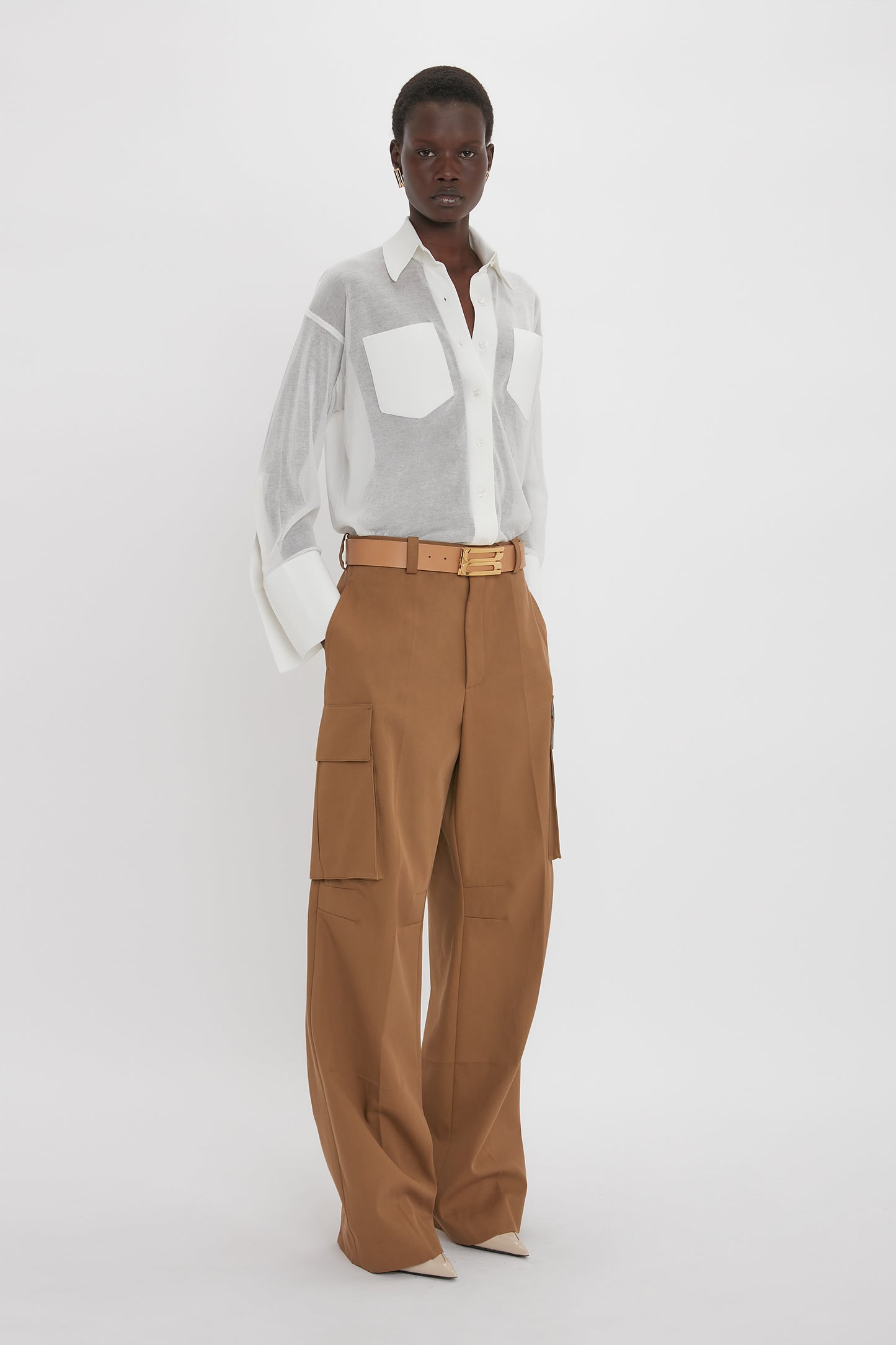 Person stands against a plain backdrop, wearing a semi-sheer Victoria Beckham Pocket Detail Shirt In White and high-waisted brown cargo pants with a beige belt, exuding casual sophistication.