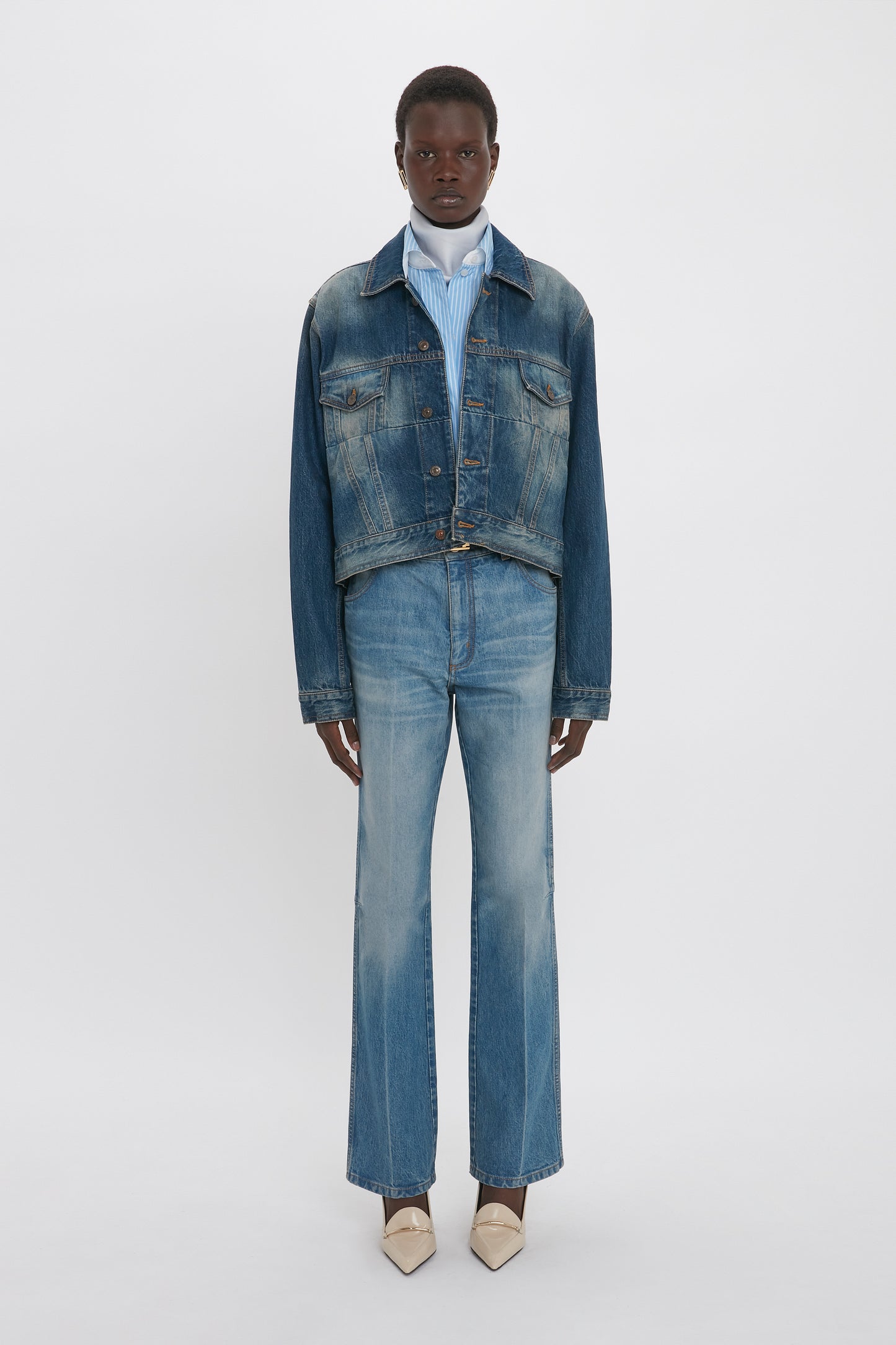 A standing person wearing a denim jacket over a blue striped shirt, paired with Relaxed Flared Jean In Broken Vintage Wash by Victoria Beckham featuring deconstructed detailing and beige pointed shoes, against a plain white background.