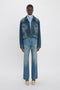 A standing person wearing a denim jacket over a blue striped shirt, paired with Relaxed Flared Jean In Broken Vintage Wash by Victoria Beckham featuring deconstructed detailing and beige pointed shoes, against a plain white background.