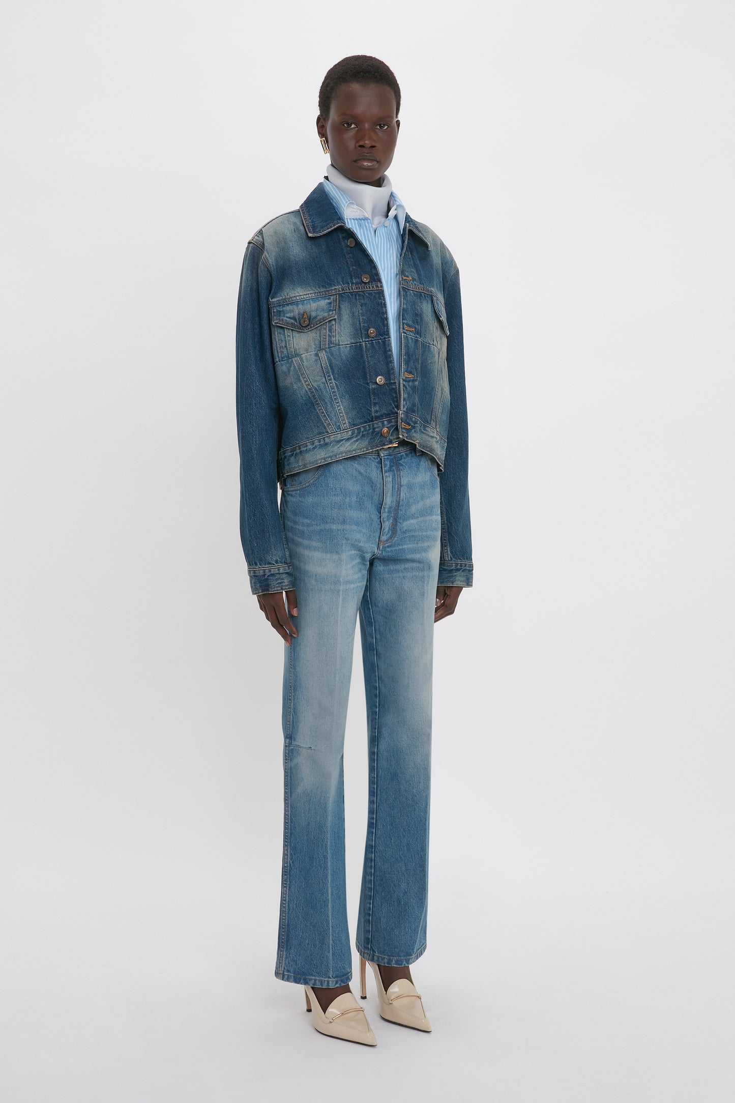 A person stands against a plain background, wearing a denim jacket with deconstructed detailing and 100% cotton Relaxed Flared Jean In Broken Vintage Wash from Victoria Beckham paired with a blue collared shirt underneath and light-colored shoes.