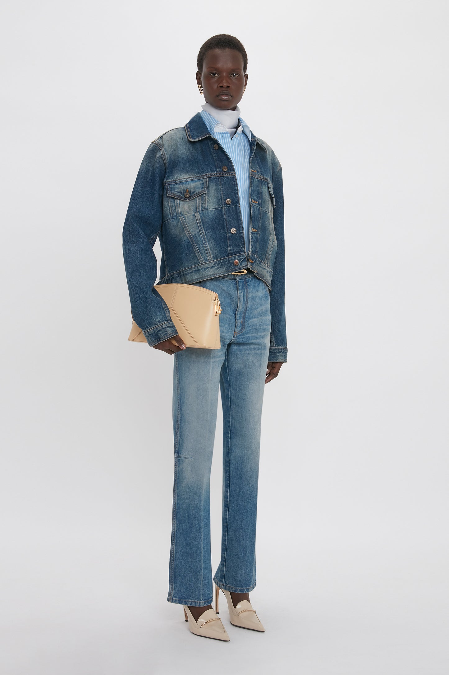 A person stands against a white background, wearing a denim jacket with a branded leather patch, Relaxed Flared Jean In Broken Vintage Wash by Victoria Beckham, a blue collared shirt, and beige shoes, holding a tan envelope clutch.