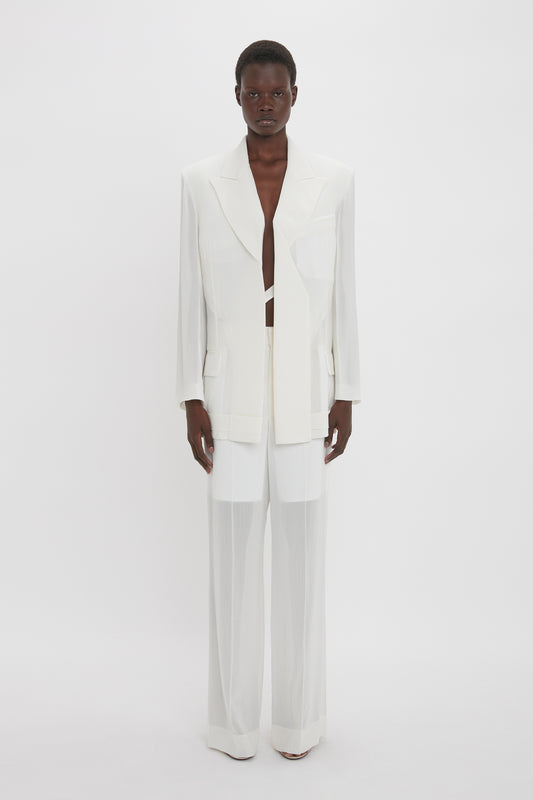 A person stands against a white background, wearing a Fold Detail Tailored Jacket In White by Victoria Beckham with a deep neckline, paired with white wide-legged pants and white heels.