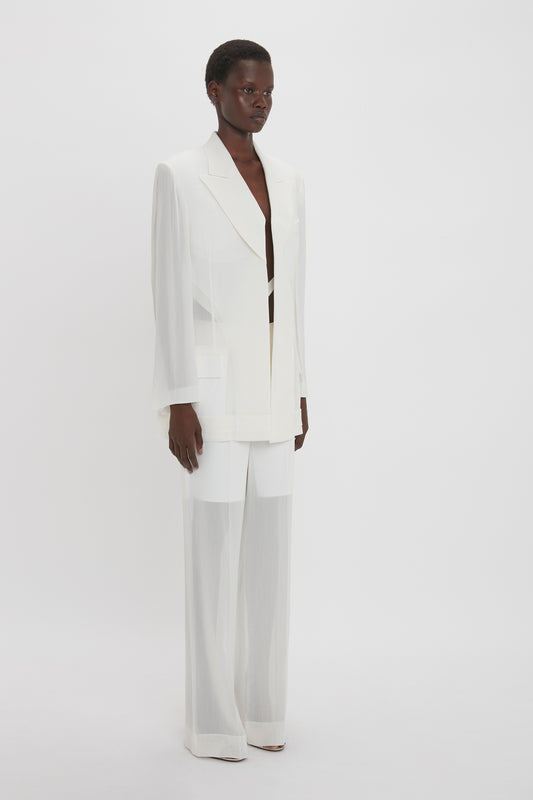 A person stands against a plain white background, wearing a white tailored suit with a double-breasted jacket and wide-leg pants. The suit, crafted from featherweight wool, boasts intricate fold detail tailored to perfection. The jacket is the Fold Detail Tailored Jacket In White by Victoria Beckham.