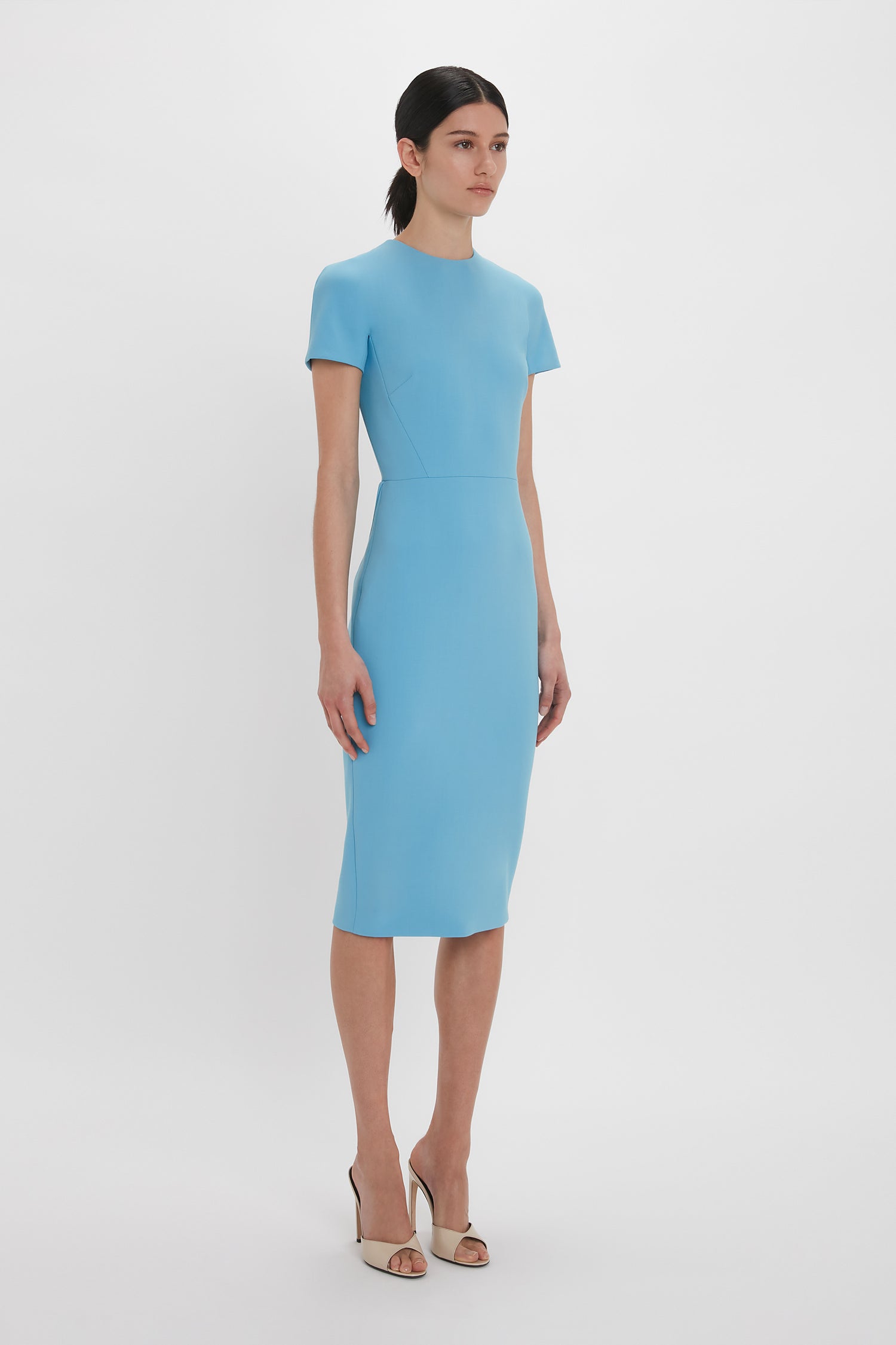 A woman in an Exclusive Fitted T-Shirt Dress In Periwinkle Blue by Victoria Beckham, paired with silver heels, standing against a white background.