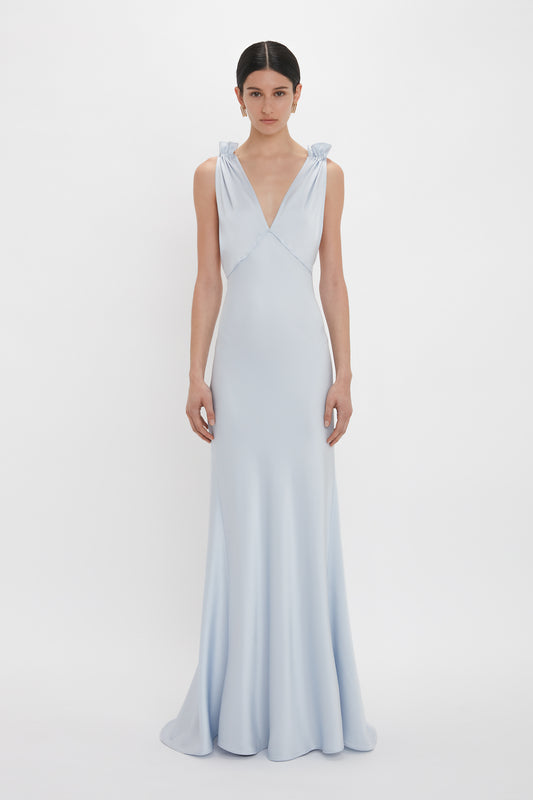 A woman is standing wearing a Victoria Beckham Exclusive Gathered Shoulder Cami Floor-Length Gown In Ice Blue with a deep V-neckline and gathered shoulder straps in a plain white background.