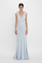 A woman is standing wearing a Victoria Beckham Exclusive Gathered Shoulder Cami Floor-Length Gown In Ice Blue with a deep V-neckline and gathered shoulder straps in a plain white background.