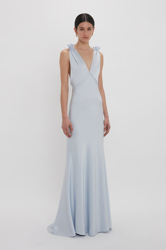 A person stands wearing an Exclusive Gathered Shoulder Cami Floor-Length Gown In Ice Blue by Victoria Beckham, made of crepe back satin, featuring a V-neck and bow accents on the shoulder straps against a plain white background.