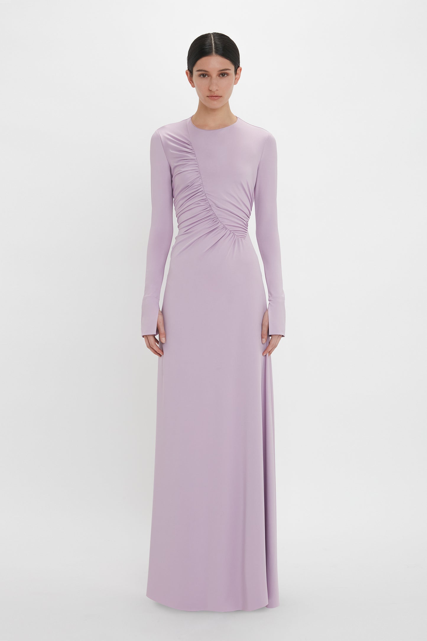 A person stands against a plain background wearing a long-sleeved, floor-length Victoria Beckham Ruched Detail Floor-Length Gown In Petunia, showcasing understated glamour with gathered detailing on one side.