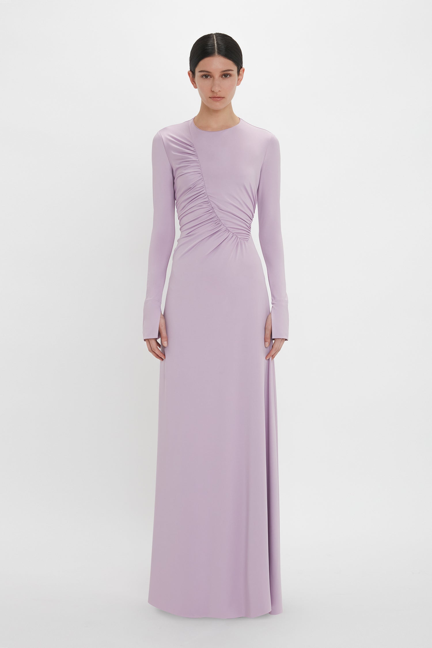 A person stands against a plain background wearing a long-sleeved, floor-length Victoria Beckham Ruched Detail Floor-Length Gown In Petunia, showcasing understated glamour with gathered detailing on one side.