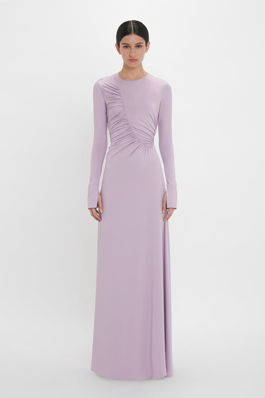 A person stands against a plain background wearing a long-sleeved, floor-length Victoria Beckham Ruched Detail Floor-Length Gown In Petunia, showcasing understated glamour with gathered detailing on one side.