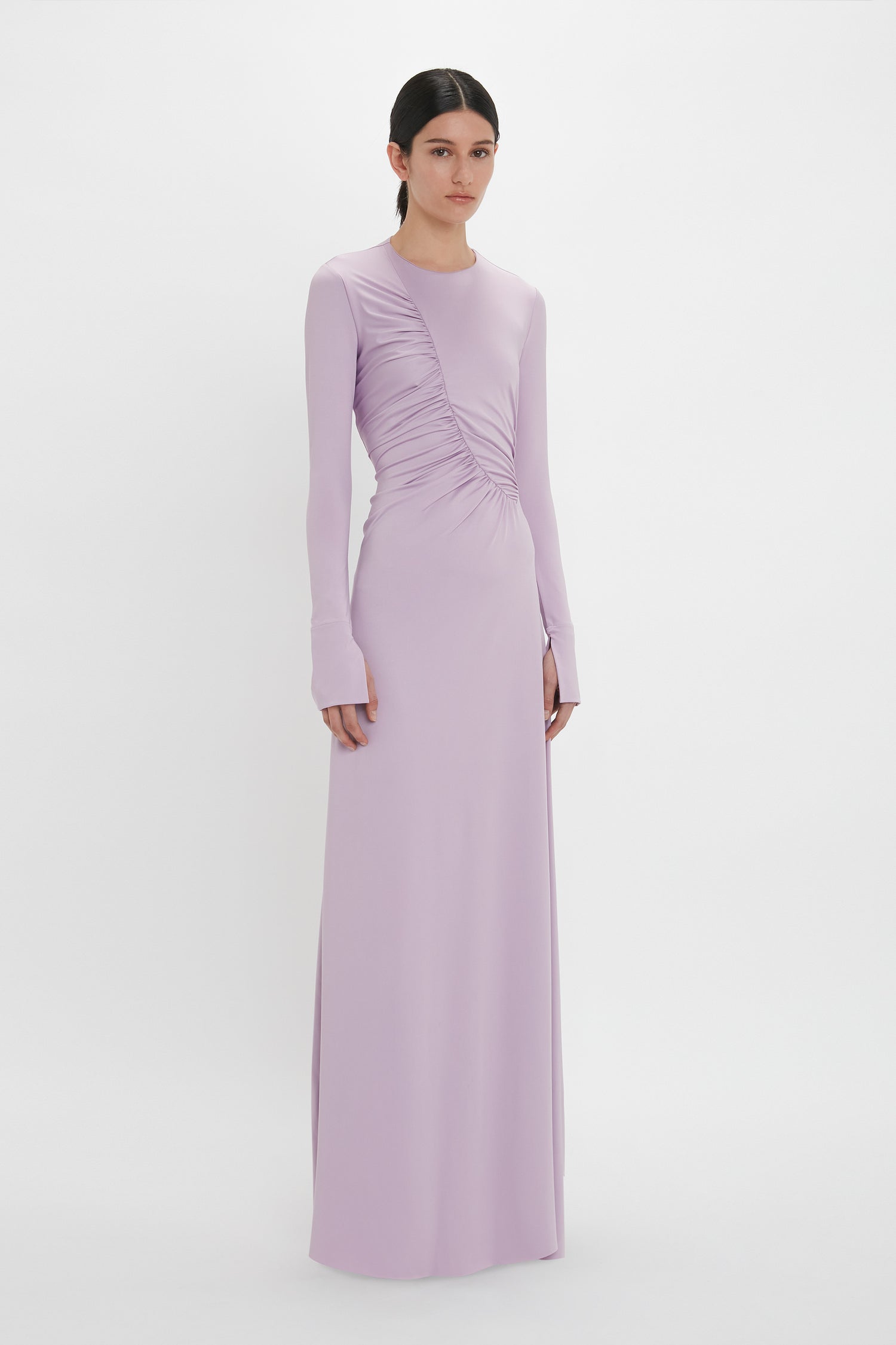 A person is standing and wearing a long-sleeved, floor-length Ruched Detail Floor-Length Gown In Petunia by Victoria Beckham that embodies understated glamour. The background is plain white.