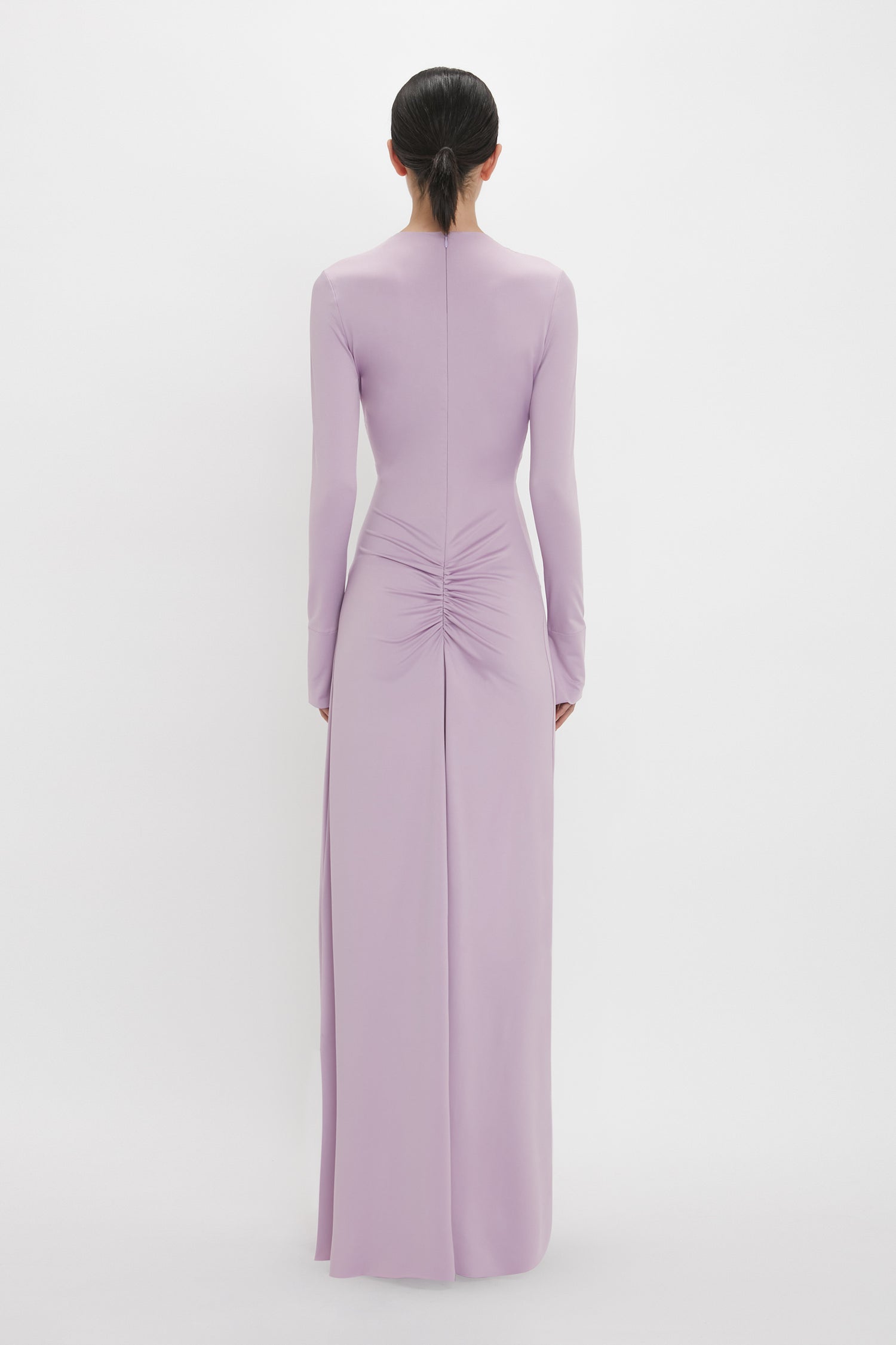 A person with dark hair in a low bun is wearing a long-sleeved, floor-length, light purple Victoria Beckham Ruched Detail Floor-Length Gown In Petunia with gathered detail at the waist, standing with their back to the camera, exuding understated glamour.