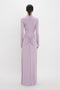 A person with dark hair in a low bun is wearing a long-sleeved, floor-length, light purple Victoria Beckham Ruched Detail Floor-Length Gown In Petunia with gathered detail at the waist, standing with their back to the camera, exuding understated glamour.