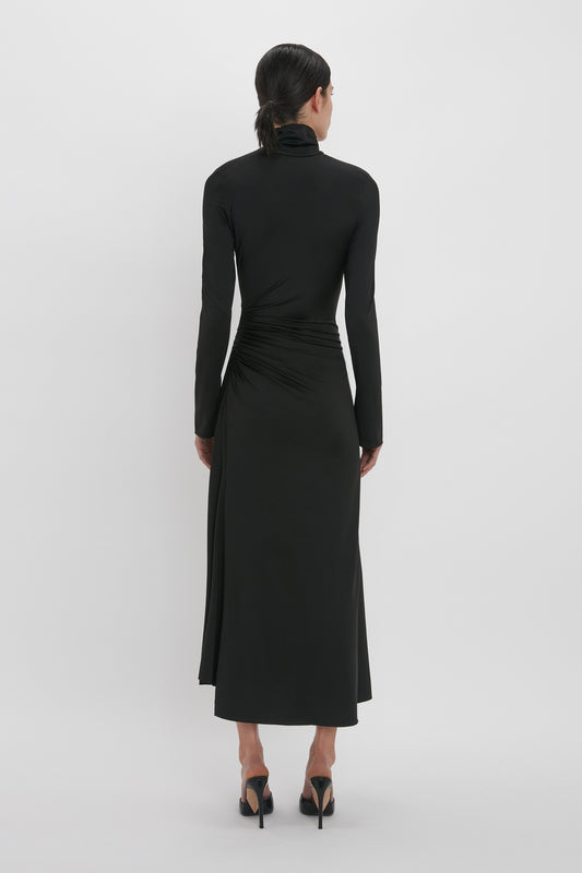 A woman viewed from behind, wearing a Victoria Beckham High Neck Asymmetric Draped Dress In Black with ruched details at the waist and high heels.