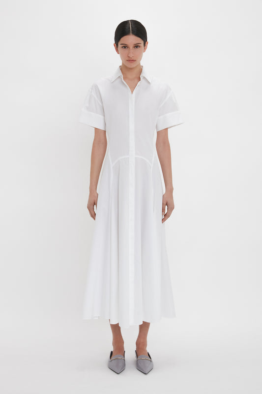 A person stands against a white background wearing a mid-length Panelled Shirt Dress In White by Victoria Beckham made from organic cotton poplin, with short sleeves and grey pointed shoes.