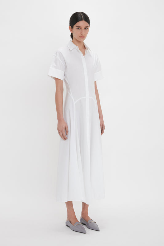 A person stands against a white background wearing the Panelled Shirt Dress In White from Victoria Beckham and grey pointed-toe flats. They have dark hair pulled back neatly.