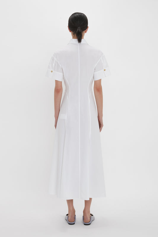 Woman stands facing away, wearing a tailored Panelled Shirt Dress In White by Victoria Beckham made from organic cotton poplin. The calf-length white dress features short sleeves and button details, set against a plain white background.