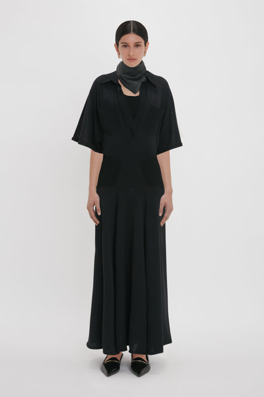 Person standing against a plain background, wearing a long, black, flowy **Panelled Knit Dress In Black** with short sleeves from the **Victoria Beckham** brand. A black scarf is around their neck, and they have dark hair tied back.