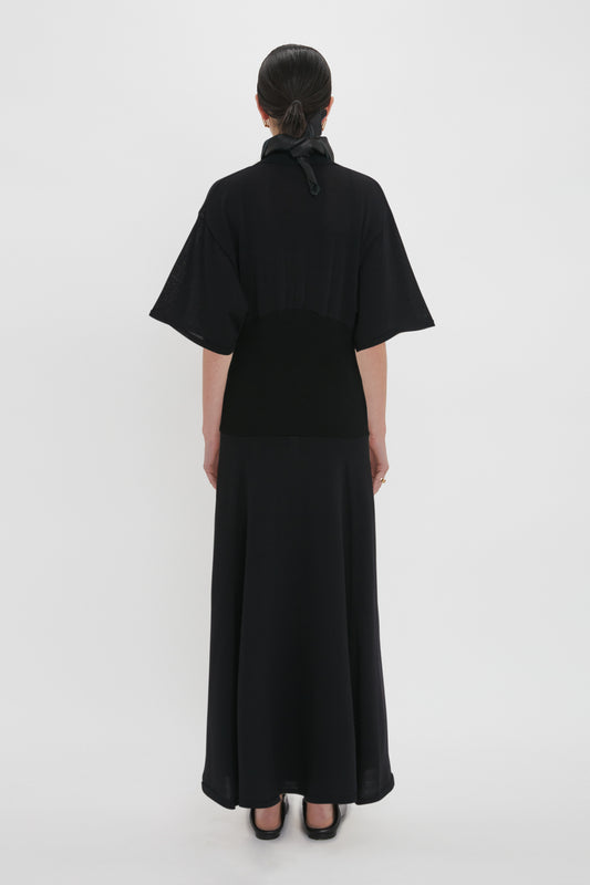 A person is seen from behind wearing an elegant black gown with elbow-length sleeves and a long skirt. The Panelled Knit Dress In Black from Victoria Beckham features a high collar with a bow at the back, embodying timeless sophistication.