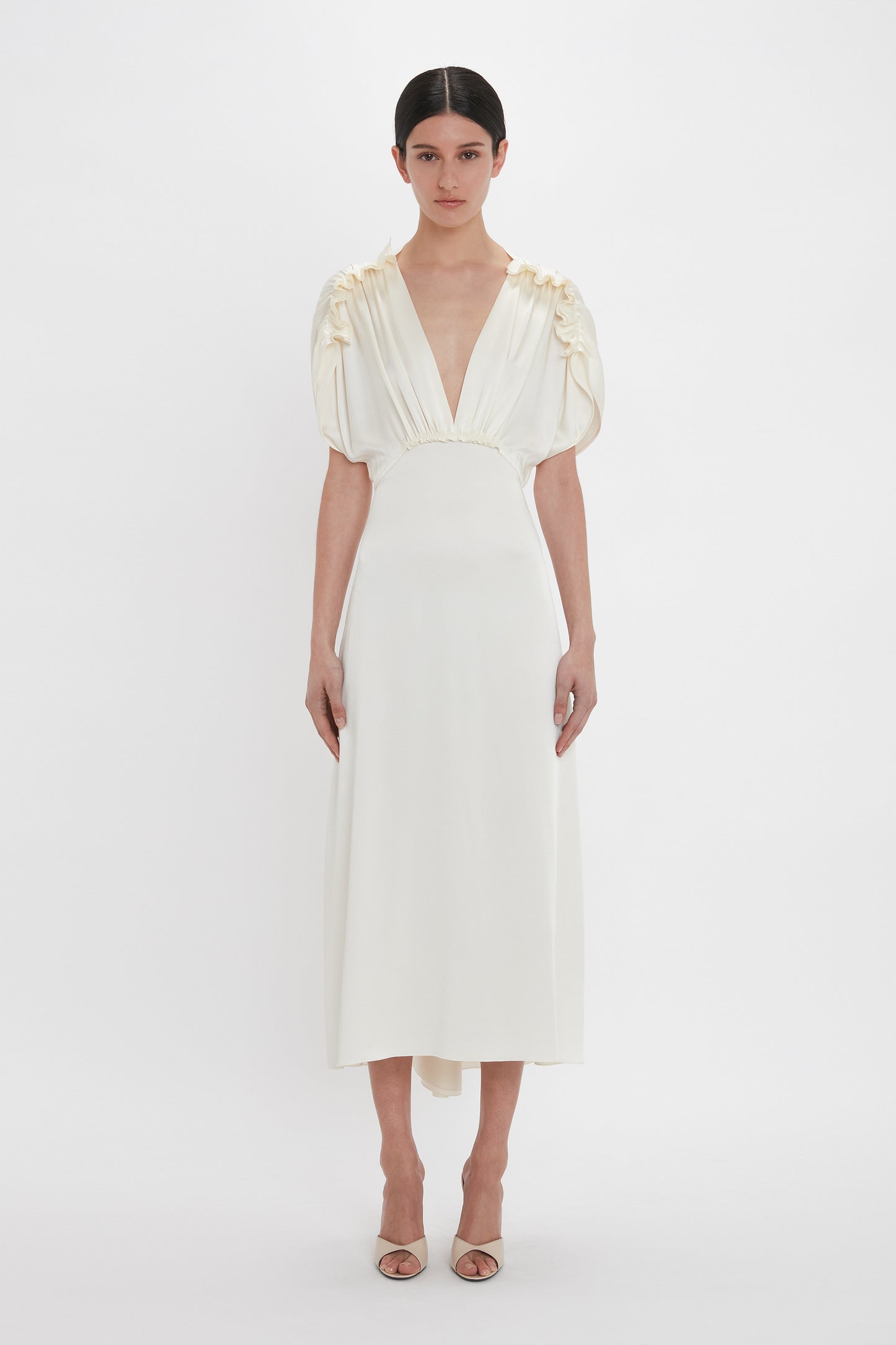 A person stands against a white background, wearing an Exclusive V-Neck Ruffle Midi Dress In Ivory by Victoria Beckham, paired elegantly with beige heeled sandals.