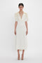 A person stands against a white background, wearing an Exclusive V-Neck Ruffle Midi Dress In Ivory by Victoria Beckham, paired elegantly with beige heeled sandals.