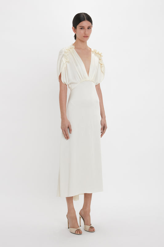A person stands against a plain background wearing a white, floor-length dress with short, ruffled sleeves. They showcase the Classic Mule In Macadamia Calf Leather by Victoria Beckham with seductive curved heels and flattering toe cleavage.