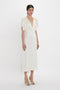 A woman stands wearing an Exclusive V-Neck Ruffle Midi Dress In Ivory by Victoria Beckham, crafted from fluid crepe back satin. The white, mid-length dress features a deep V-neckline and short puffed sleeves. She pairs the elegant outfit with open-toe high heels.