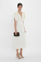 A person stands against a plain background, adorned in a Victoria Beckham Exclusive V-Neck Ruffle Midi Dress In Ivory made from fluid crepe back satin. They're holding a black clutch and wearing white high-heeled shoes.