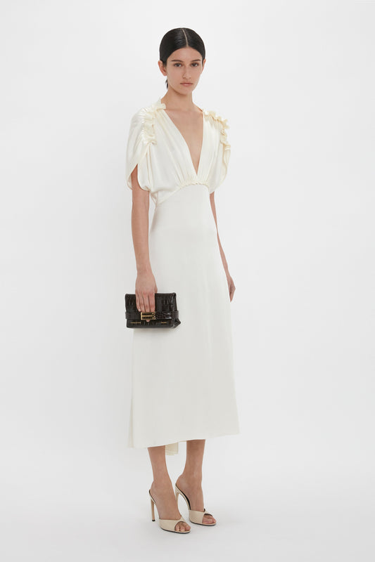 A person wearing an elegant, white, mid-length dress with a deep V-neck and ruffled shoulders, holding a Victoria Beckham Mini B Pouch In Croc Effect Espresso Leather and standing against a plain white background.