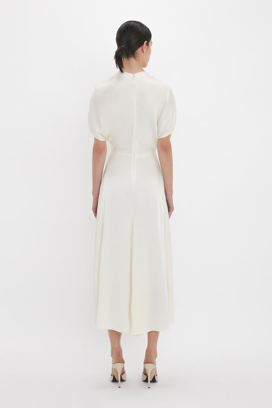 A woman with dark hair is standing with her back to the camera, showcasing an Exclusive V-Neck Ruffle Midi Dress In Ivory by Victoria Beckham. The mid-calf length dress has short sleeves and beige high-heeled shoes, set against a plain white background.