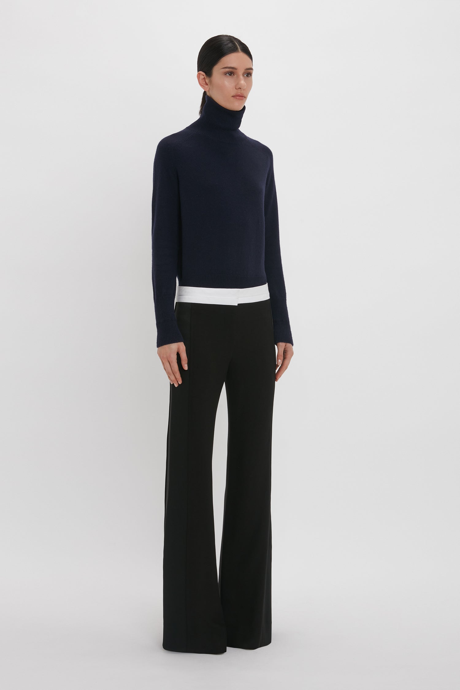 A person stands facing slightly left, donning a navy turtleneck sweater and Victoria Beckham's Side Panel Trouser In Black with a wide-leg design, complemented by a white undershirt. The backdrop is plain white.