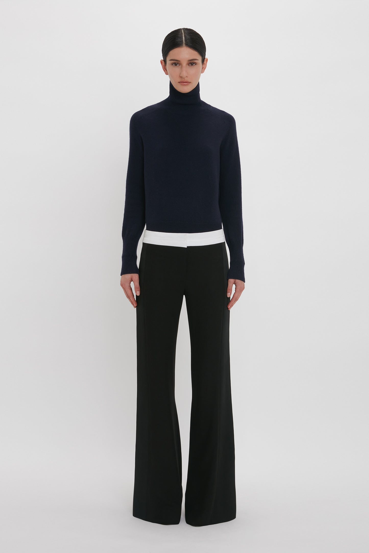 A person with dark hair wears a black turtleneck sweater and Victoria Beckham Side Panel Trouser In Black, standing against a plain white background.