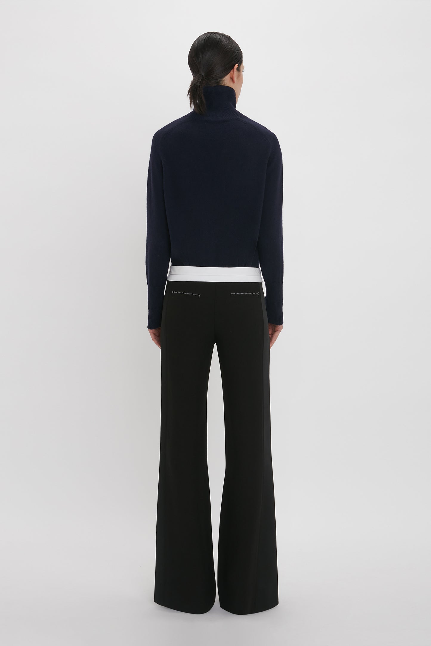 A person with dark hair stands facing away, wearing a black turtleneck, white shirt underneath, and Victoria Beckham's Side Panel Trouser In Black with a low-rise waistline. The background is plain white.