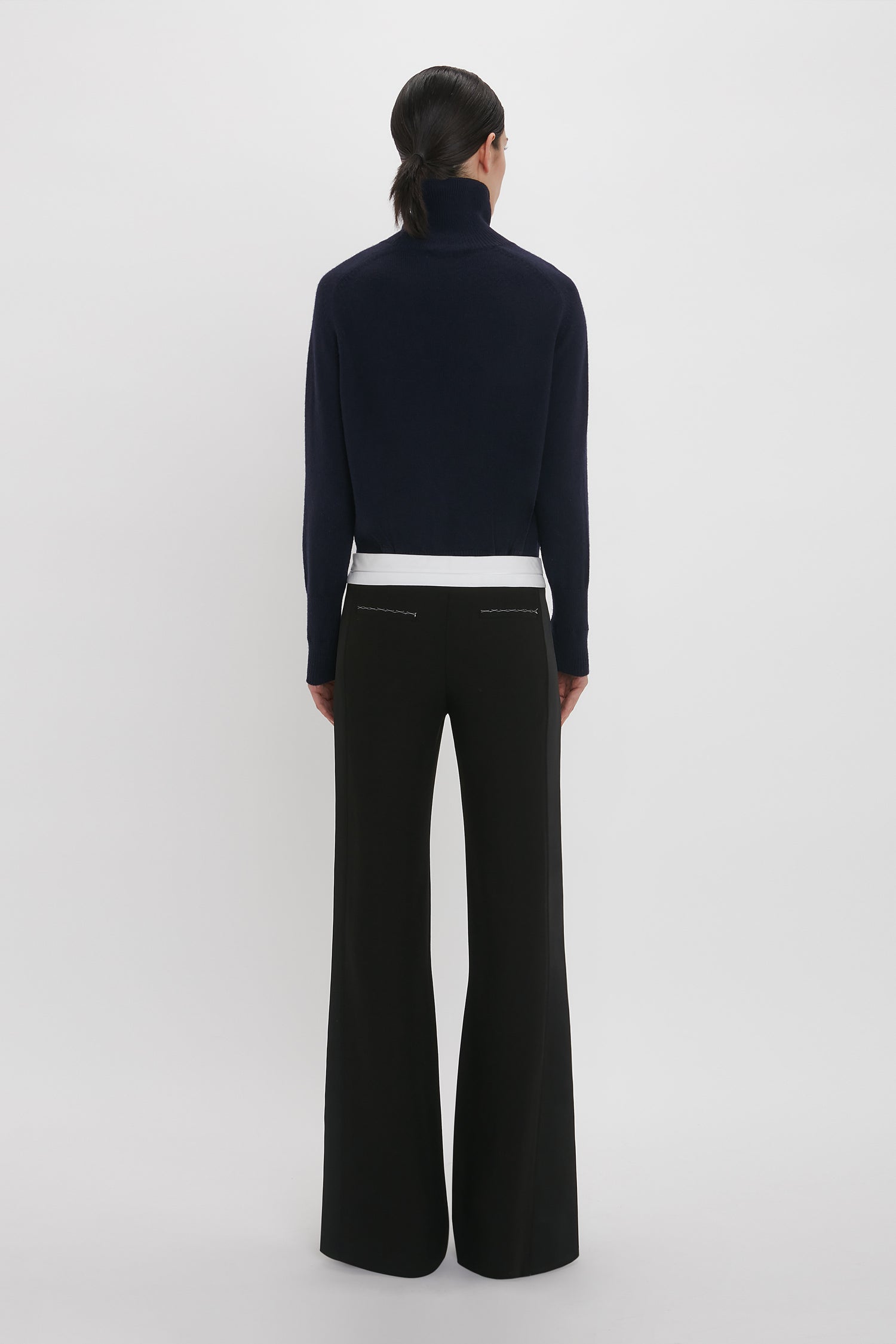 A person with dark hair stands facing away, wearing a black turtleneck, white shirt underneath, and Victoria Beckham's Side Panel Trouser In Black with a low-rise waistline. The background is plain white.