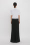 Woman in a white t-shirt and Victoria Beckham black floor-length box pleat skirt, standing against a plain white background.