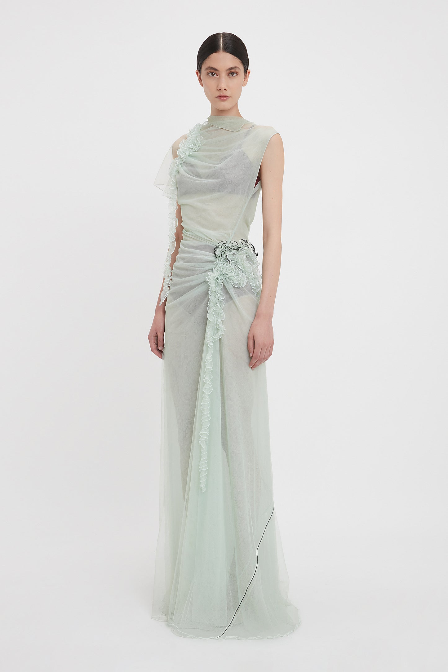 A model stands wearing the Victoria Beckham Gathered Tulle Detail Floor-Length Dress In Jade, crafted from delicate sea foam green tulle with a sheer, flowing design and floral ruche detailing on the side.