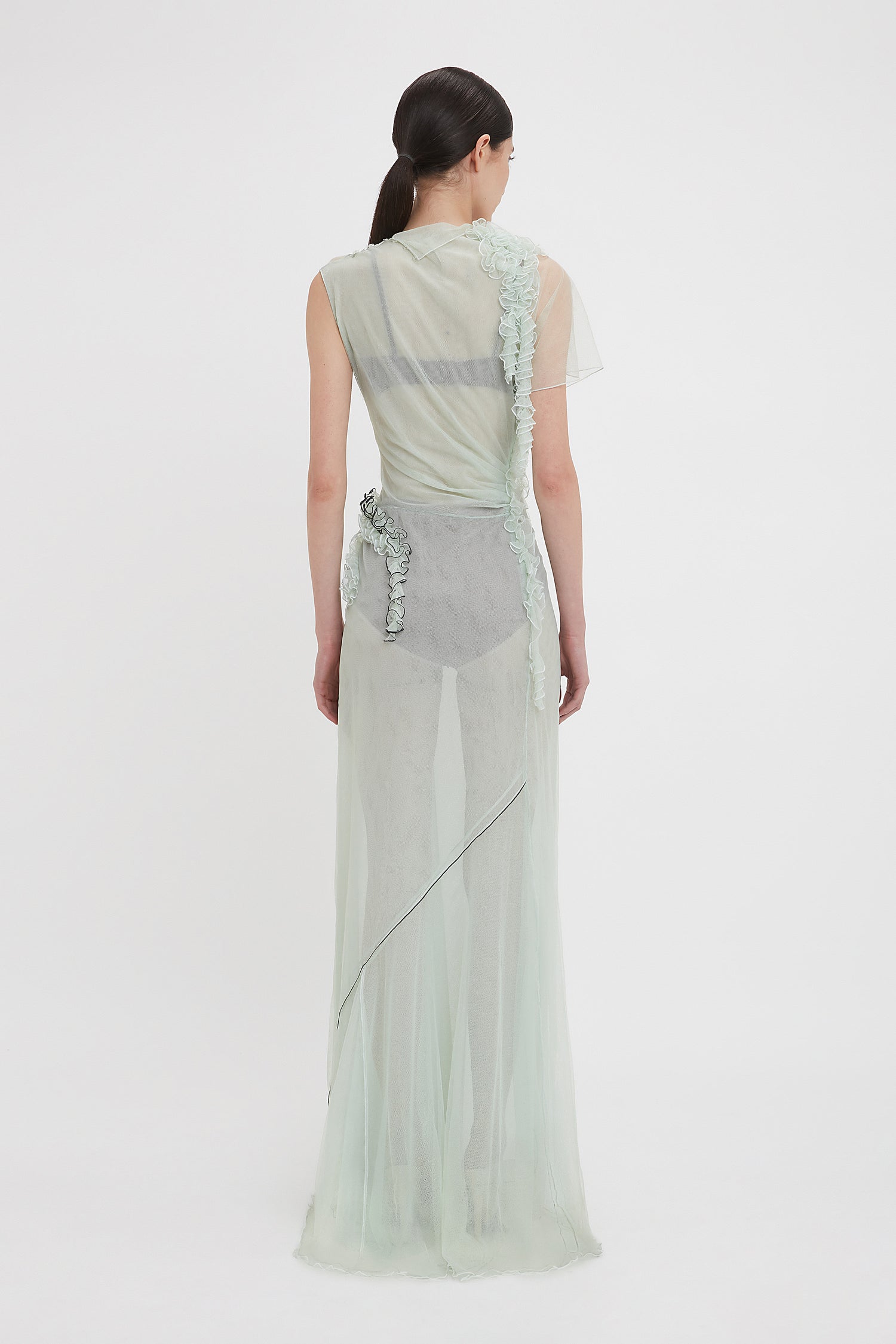 A woman with dark hair stands facing away, wearing the Victoria Beckham Gathered Tulle Detail Floor-Length Dress In Jade, adorned with floral embellishments and an asymmetrical hemline.