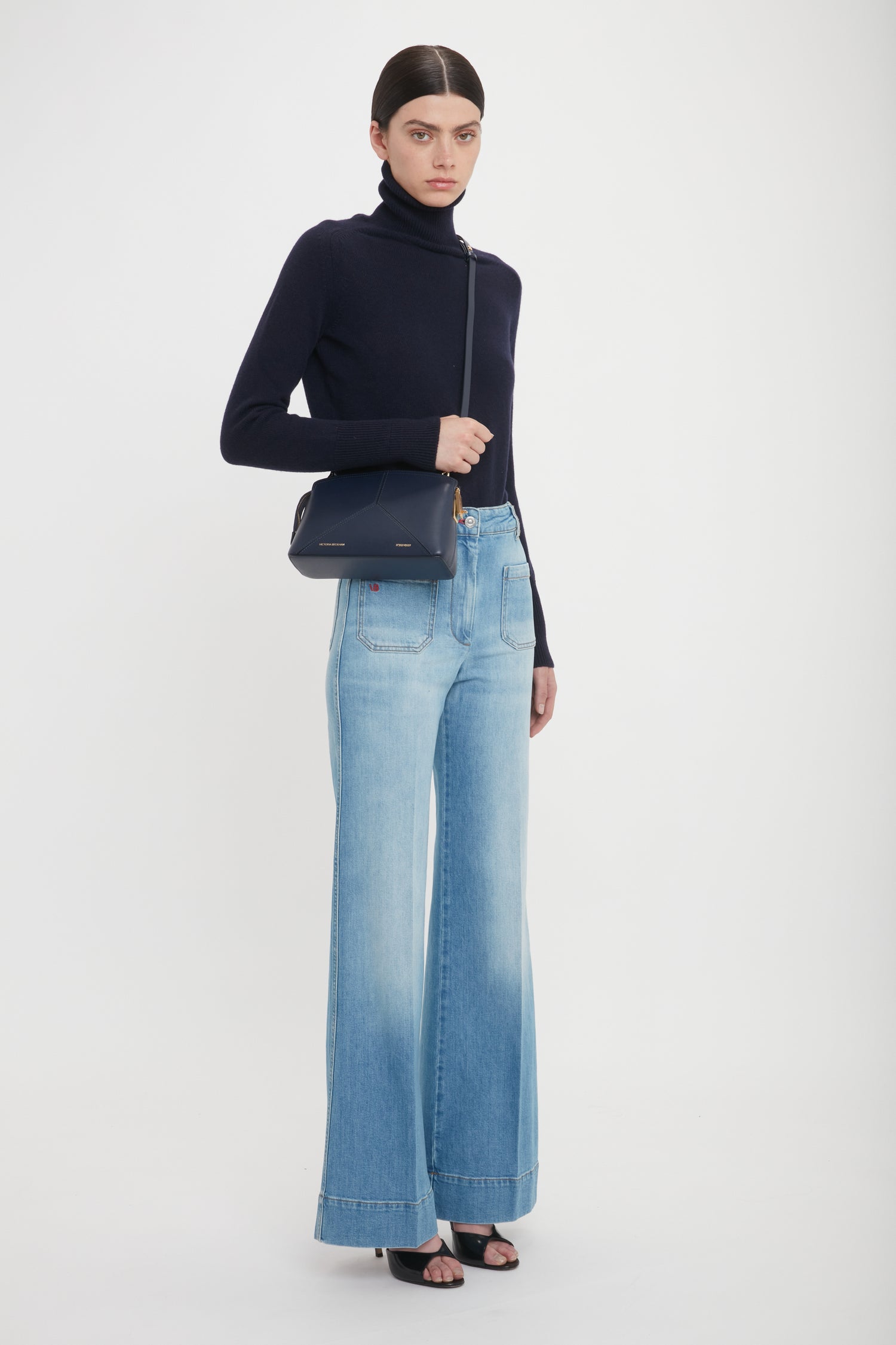 A person stands against a plain background wearing a black turtleneck, light blue wide-leg jeans, and black open-toe heels, holding the Exclusive Victoria Crossbody Bag In Navy Leather by Victoria Beckham over the shoulder.