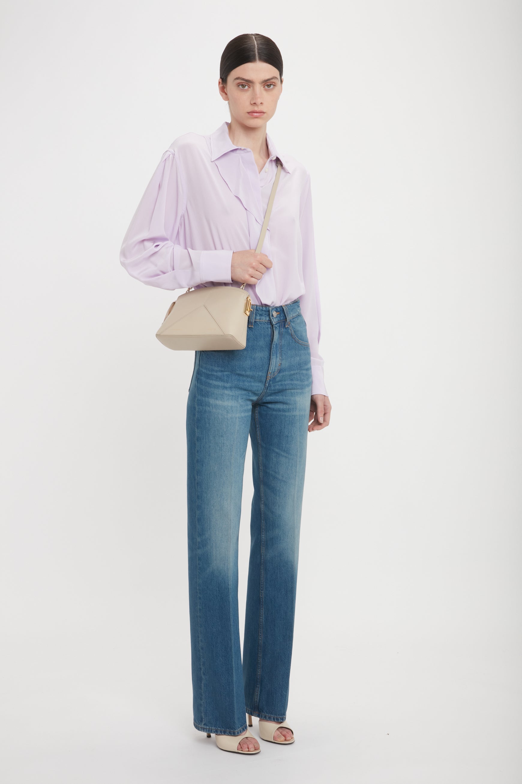 A person with long, dark hair in a middle part wears a light lavender blouse, high-waisted blue jeans, beige sandals, and carries a Victoria Beckham Victoria Crossbody Bag In Taupe Leather with an adjustable and removable strap, standing against a plain white background.