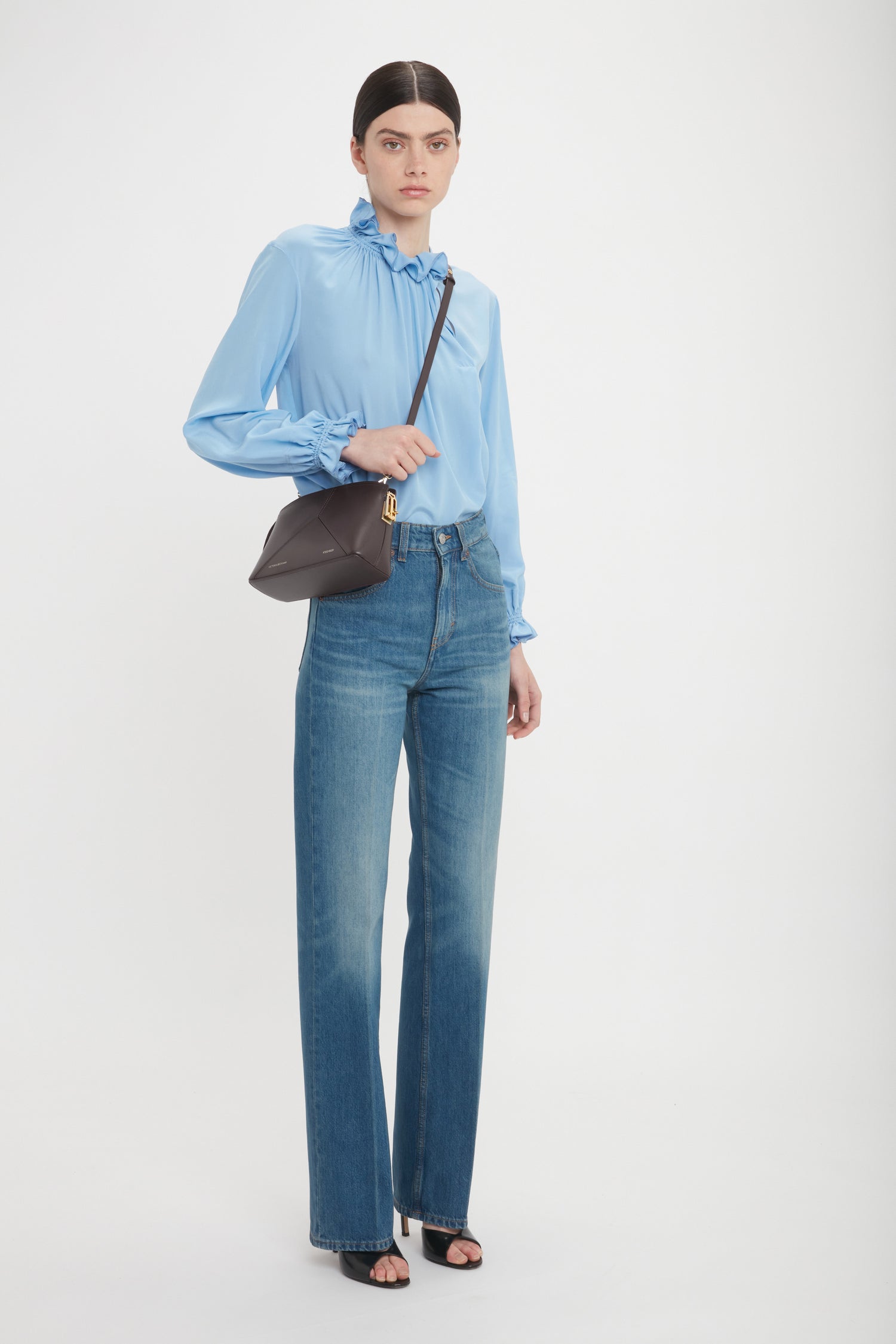 A person wearing a light blue blouse, blue jeans, black open-toe heels, and an Exclusive Victoria Crossbody Bag In Brown Leather by Victoria Beckham with its signature V shape in elegant calf leather stands against a plain white background.