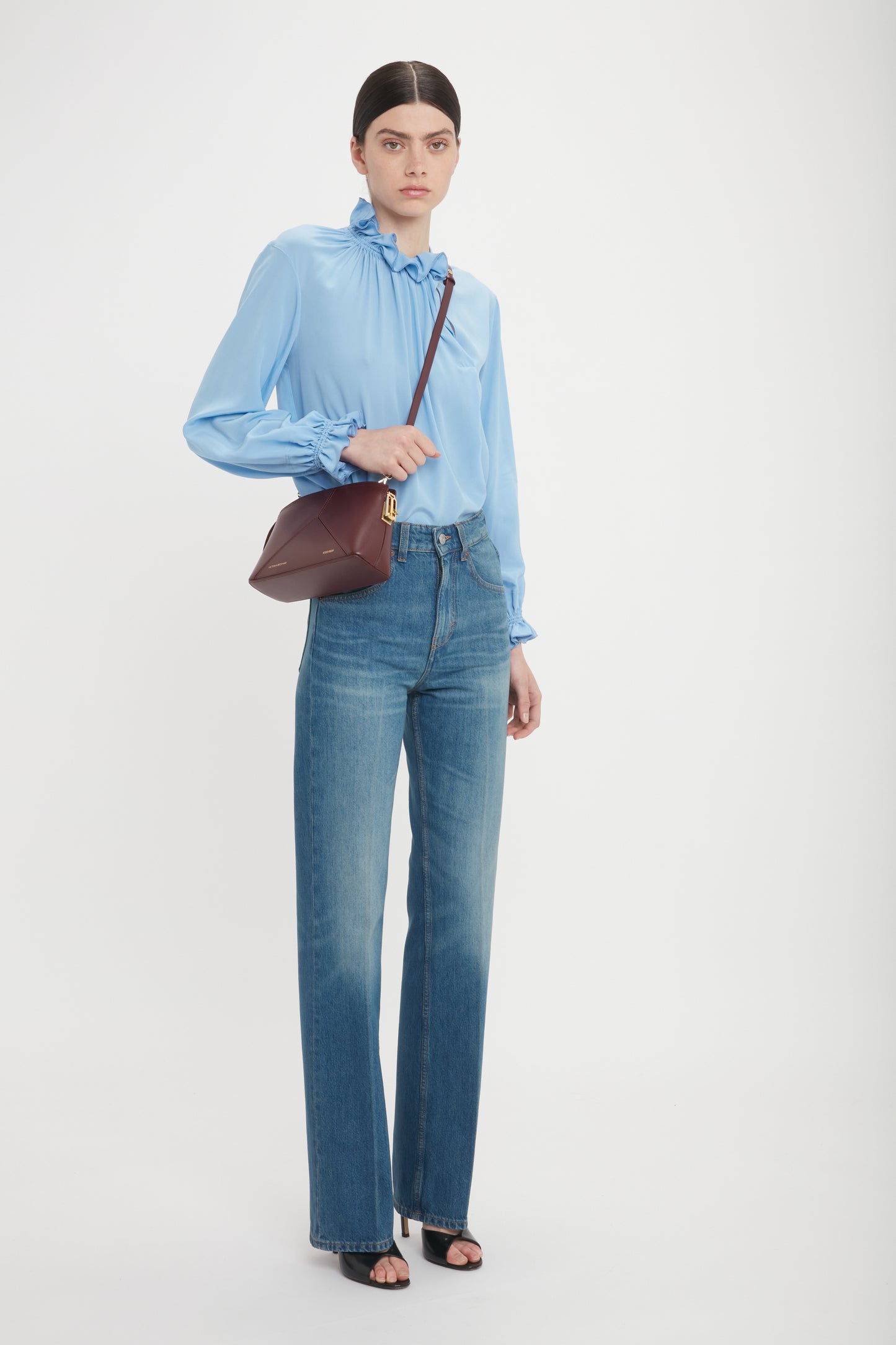 Person standing against a white background, wearing a light blue long-sleeve blouse, high-waisted blue jeans, black open-toe heels, and holding the Victoria Crossbody Bag In Burgundy Leather by Victoria Beckham.