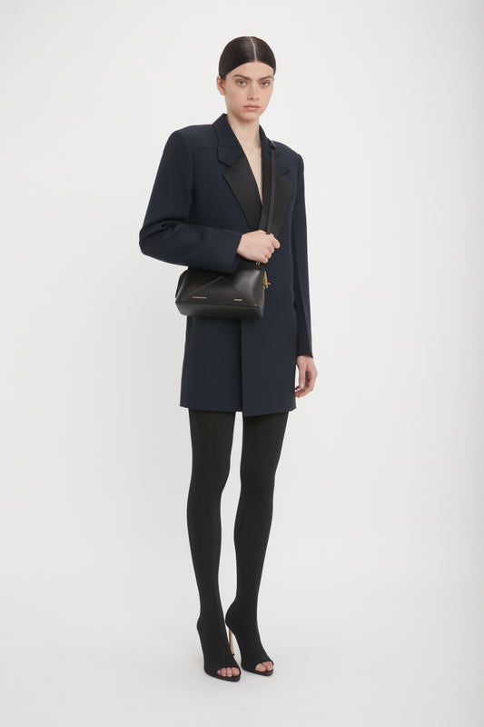 A person stands against a plain background wearing a dark blazer, black tights, open-toe heels, and carrying the Victoria Beckham Victoria Crossbody Bag In Black Leather.
