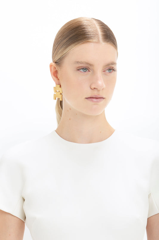 A woman with slicked back hair wearing a white top and a large geometric gold earring, facing forward against a white background, holds a Victoria Beckham Fitted T-shirt Dress In Ivory.